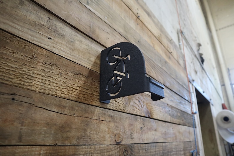 Puede incluir: Portabicicletas de metal negro montado en una pared de madera. El soporte tiene forma de bicicleta con un dise&ntilde;o recortado.
