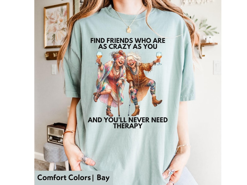 Puede incluir: Camiseta azul claro con el texto "FIND FRIENDS WHO ARE AS CRAZY AS YOU AND YOU'LL NEVER NEED THERAPY". La camiseta presenta un gr&aacute;fico de dos mujeres mayores sosteniendo copas de vino. La camiseta es de material Comfort Colors.