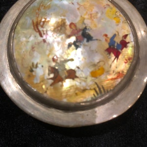 May include: A silver circular pendant with a round, iridescent mother-of-pearl inlay depicting a scene of people riding horses in a landscape.