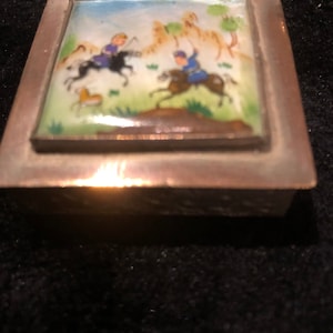 Puede incluir: Una pequeña caja cuadrada de plata con una tapa con bisagras. La tapa presenta una escena pintada de dos personas montando a caballo en un paisaje con árboles y colinas. La caja está decorada con un patrón floral.