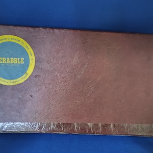 May include: A brown cardboard box with a yellow circle sticker that says "SCRABBLE" with the words "Manufactured by SELCHOW & RIGHTER CO., NEW YORK, N.Y." around the circle.