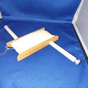 May include: A wooden kite spool with white string, featuring a rectangular wooden frame and two white handles. The spool is designed for holding and dispensing kite string. The background is a solid blue.