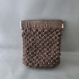 May include: A small, brown macrame pouch with a rectangular shape. The pouch features a textured, knotted design and a reinforced top edge with metal accents. The overall aesthetic is handmade and rustic.