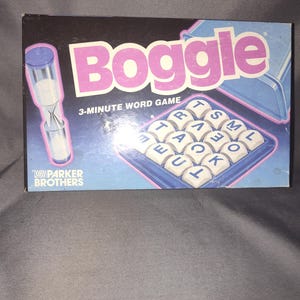 May include: A vintage Boggle word game box with a black background and pink lettering. The box features a 3-minute timer and a grid of lettered dice. The Parker Brothers logo is visible.