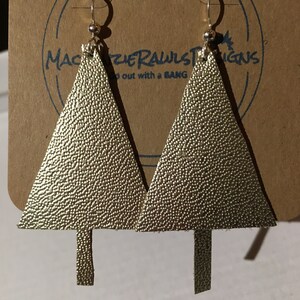 May include: A pair of gold-colored leather earrings shaped like Christmas trees. The earrings have a textured surface and are hanging from silver hooks.