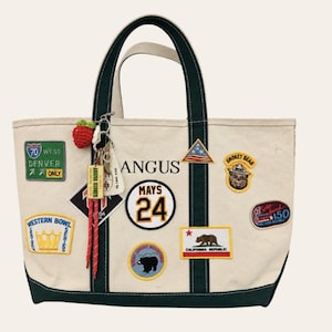 May include: A cream-colored canvas tote bag with dark green trim and handles, decorated with various embroidered patches including a Denver road sign, Smokey Bear, California Republic flag, and the text "ANGUS" and "MAYS 24". A small crocheted strawberry charm hangs from the handle.