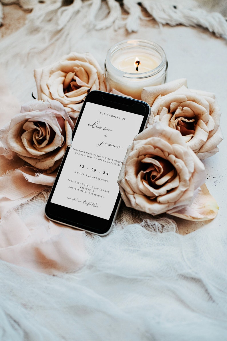 May include: A wedding invitation on a smartphone screen with the text "The Wedding of Olivia + Jason" and details about the ceremony and reception. The phone is surrounded by beige roses and a lit candle.