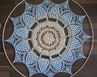 Puede incluir: Un atrapasueños circular hecho a mano con un aro de metal blanco. El intrincado diseño de ganchillo presenta un degradado de colores, que transita de un centro beige claro a tonos de azul. El diseño tiene un patrón de estrella.