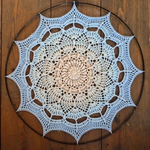 Può includere: Una decorazione da parete circolare con un mandala all'uncinetto caratterizzato da una sfumatura di colore che passa da un centro color pesca tenue a un bordo esterno azzurro, fissato a un sottile cerchio in metallo nero con dodici punte.