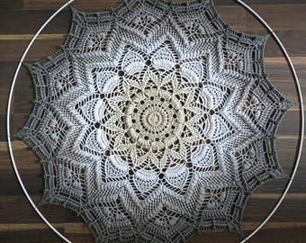May include: A handmade, crocheted mandala wall hanging. The circular design features a gradient of colors, transitioning from a light beige center to shades of gray and white, framed by a white hoop. The intricate pattern showcases detailed stitch work.