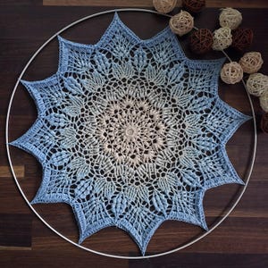 May include: A circular crochet mandala featuring a gradient color pattern that transitions from a cream center to a light blue outer edge. The piece is mounted within a thin white circular frame and is surrounded by small decorative rattan spheres.