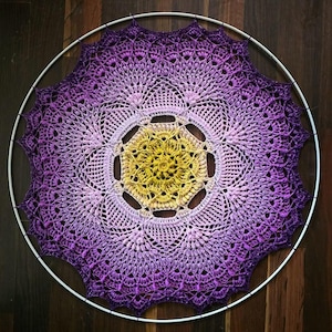 May include: A handcrafted, circular wall hanging featuring a crocheted design in shades of purple, lavender, and yellow. The intricate pattern is set within a white metal hoop, creating a decorative piece for home decor.
