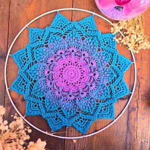 May include: A circular crochet mandala wall hanging featuring a vibrant gradient of colors transitioning from a bright purple center to a deep teal outer edge, securely attached to a thin white metal hoop frame with delicate yarn stitching.