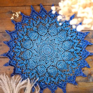 Może przedstawiać: Ręcznie robiona, okrągła serwetka w odcieniach niebieskiego. Złożony, ażurowy wzór ma centralny okrągły wzór, który promieniuje na zewnątrz, z falistą krawędzią. Kolor przechodzi od jasnoniebieskiego do ciemnoniebieskiego.
