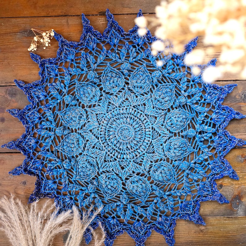 K&ouml;nnte beinhalten: Ein handgefertigtes, rundes Deckchen in Blaut&ouml;nen. Das detaillierte, offene Design weist ein zentrales kreisf&ouml;rmiges Muster auf, das sich nach au&szlig;en ausdehnt, mit einem gewellten Rand. Die Farbe wechselt von hell- zu dunkelblau.