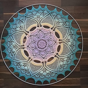 May include: A circular, hand-crocheted mandala in a gradient of colours, from blue and green to yellow, pink, and purple. The detailed design incorporates floral and geometric motifs, set within a white metal hoop. The mandala is presented on a dark wooden surface.