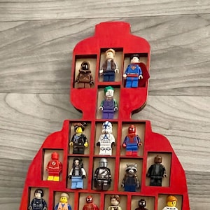 Puede incluir: Un expositor de madera rojo para minifiguras Lego con 36 ranuras, lleno de diversas minifiguras Lego, incluyendo superhéroes, personajes de Star Wars y otras figuras populares. El expositor tiene forma de minifigura Lego y tiene un recorte para la cabeza y los brazos.