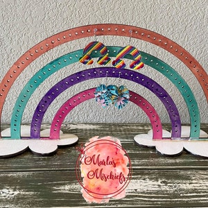 May include: A rainbow-coloured wooden earring stand with a cloud-shaped base. The stand has four tiers of holes for displaying earrings. The stand is decorated with a rainbow pattern and the words "Marla's Mischiefs".
