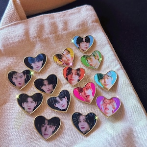 May include: A collection of 12 heart-shaped pins featuring different portraits of people. The pins are gold-colored and have a shiny finish. The portraits are in various colors and styles.