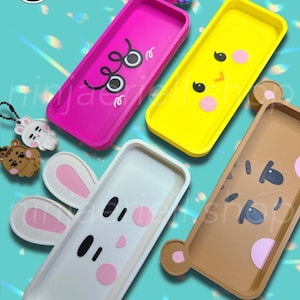 Puede incluir: Una colección de fundas de teléfono coloridas con diseños de animales de dibujos animados. Las fundas son rosas, amarillas, blancas y marrones, cada una con una cara diferente. La imagen también incluye llaveros con diseños de animales similares. El nombre de la tienda "ninjaeriell shop" es visible.