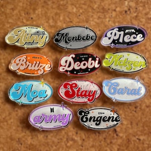 May include: Twelve colorful, round, plastic pins with the names of different K-pop groups written in a retro font. The names are: Ateez, Monbebe, P1ece, Brize, Deobi,  Netzzen, Moa, Stay, Carat, Army, Engene.
