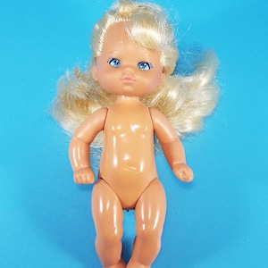 May include: A vintage plastic baby doll with blonde hair and blue eyes. The doll is nude and has a light skin tone. The doll is lying on a solid blue background. The doll's arms and legs are slightly bent.