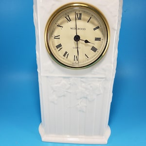 Könnte beinhalten: Eine weiße Keramikuhr mit goldfarbenem Rahmen und cremefarbenem Zifferblatt. Das Zifferblatt zeigt römische Ziffern und den Markennamen "WEDGWOOD". Die Uhr hat eine rechteckige Form mit geprägten Blattmotiven.