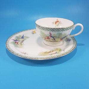 May include: A white teacup and saucer set with a decorative bird and floral design. The cup and saucer have a green and gold trim. The set is on a blue background.