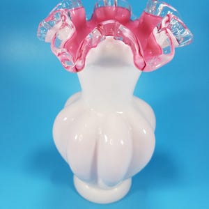Può includere: Un vaso di vetro bianco con un bordo increspato, rosa e trasparente. Il vaso ha un corpo bulboso con una serie di lobi arrotondati. Il vaso poggia su una piccola base circolare. Lo sfondo è blu unito.
