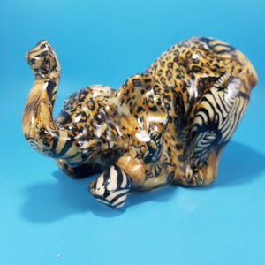 May include: A ceramic elephant figurine featuring a glossy finish with a multi-patterned surface. The body displays a mix of black and brown leopard spots, zebra stripes, and tiger-like markings across its trunk, ears, and curved back.