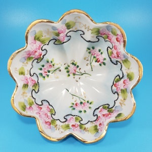 May include: A white ceramic serving dish with a scalloped edge and gold trim. The dish is decorated with pink floral designs and black accents. The dish is approximately 10 inches in diameter.