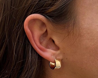 Puede incluir: Pendientes de aro de oro con una banda gruesa y redondeada. Los pendientes se llevan en la oreja de una persona.