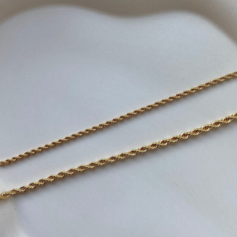 May include: Two gold-colored rope chains of different thicknesses are displayed against a white background. The chains have a twisted design, reflecting light. The jewelry is likely for necklaces or bracelets.