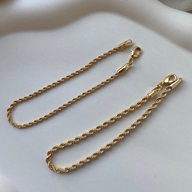 May include: Two gold-colored rope chain bracelets with lobster claw clasps. The bracelets are lying on a white surface. The chains have a twisted rope design and are made of a shiny, metallic material.