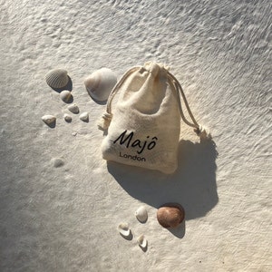 May include: A beige drawstring bag with the text "Maj&ocirc; London" in black, surrounded by various seashells. The shells vary in size and color, with shades of white, beige, and brown. The bag is made of a natural fabric.