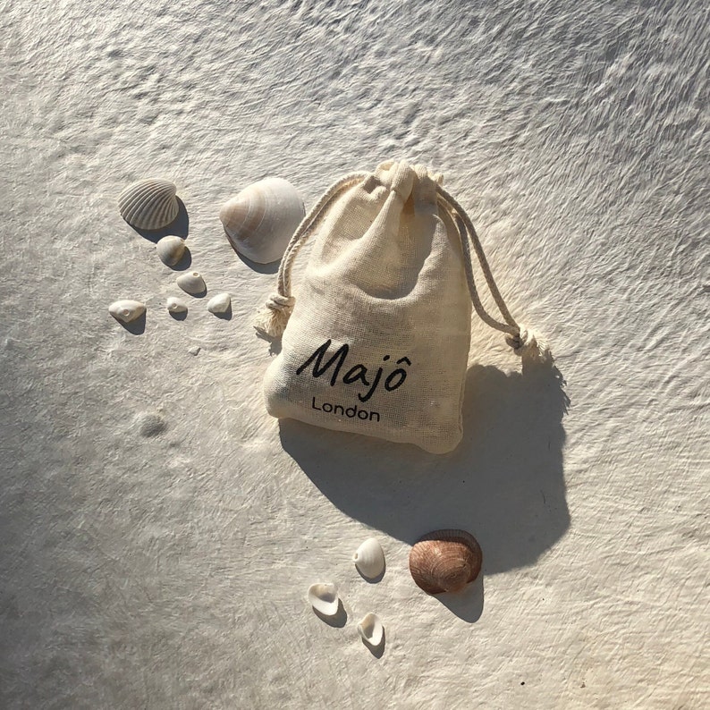 May include: A beige drawstring bag with the text "Maj&ocirc; London" in black, surrounded by various seashells. The shells vary in size and color, with shades of white, beige, and brown. The bag is made of a natural fabric.