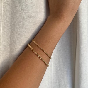 May include: Two gold-colored rope-style bracelets are worn on an arm. The bracelets are made of a twisted metal design. The arm is against a white fabric background. The bracelets are a simple, elegant accessory.