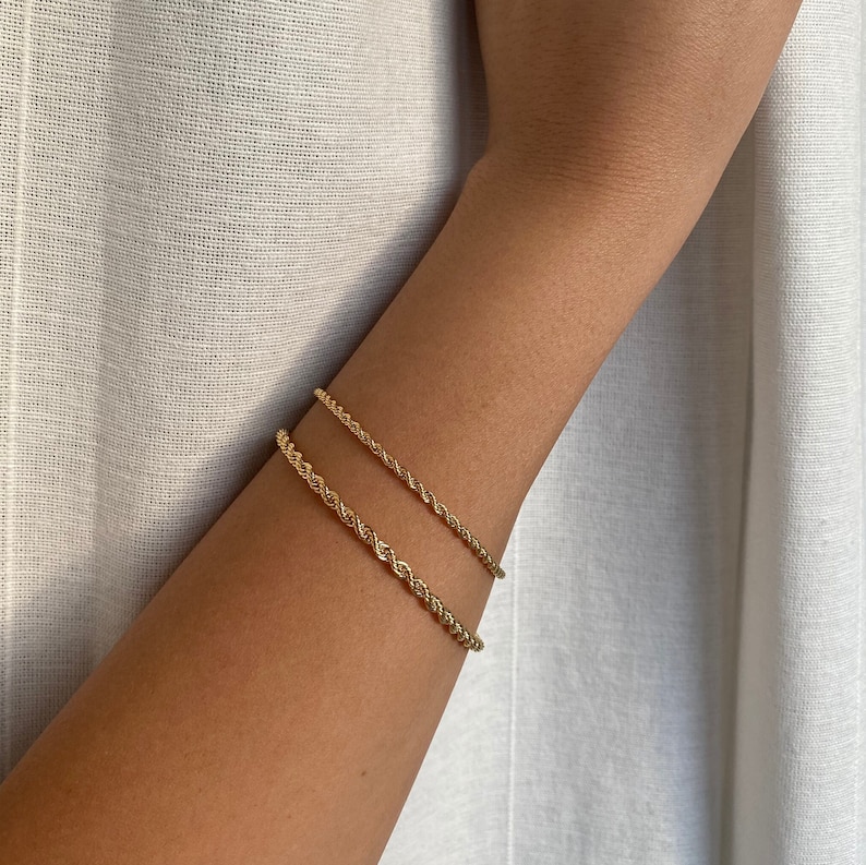 May include: Two gold-colored rope-style bracelets are worn on an arm. The bracelets are made of a twisted metal design. The arm is against a white fabric background. The bracelets are a simple, elegant accessory.