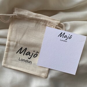 May include: A small, beige drawstring bag and a white card with the text "Maj&ocirc; London" in black script. The bag is made of a natural fabric, and the card is blank, with a small notch at the top.