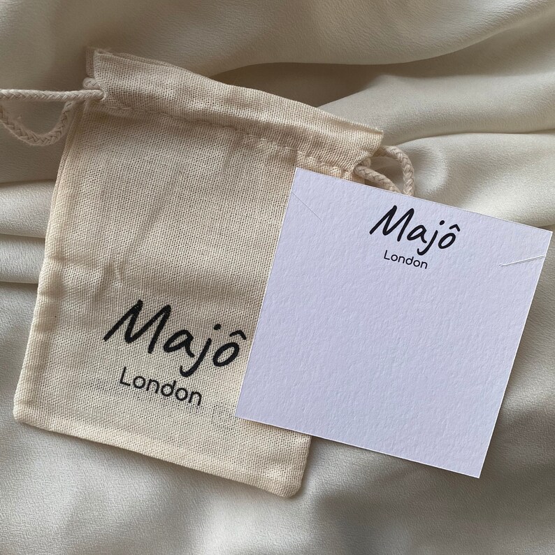 May include: A small, beige drawstring bag and a white card with the text "Maj&ocirc; London" in black script. The bag is made of a natural fabric, and the card is blank, with a small notch at the top.