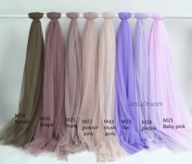 May include: Nine different colors of tulle fabric swatches hanging from a white rod. The colors are khaki, grape, nude, pinkish pink, blush pink, lilac, purple, and baby pink. The swatches are labeled with their corresponding color names and product codes.