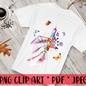 May include: A white t-shirt with a watercolor design of a hand holding a syringe with flowers and butterflies. The design is in shades of pink, blue, and green.