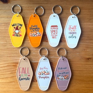 Op de afbeelding: Een verzameling van acht retro-stijl sleutelhangers in verschillende kleuren, met herfstthema's en tekst. De sleutelhangers hebben metalen ringen en bevatten zinnen als "Welcome Fall" en "Pumpkin Spice and everything nice."