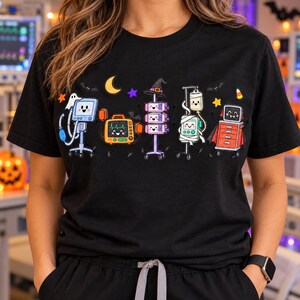 Könnte beinhalten: Ein schwarzes T-Shirt mit einem Grafikdruck von fünf anthropomorphen medizinischen Geräten mit lächelnden Gesichtern. Das Design umfasst einen Monitor, ein Herzfrequenzmessgerät, eine Pumpe, einen Infusionstropf und einen Wagen, verziert mit Halloween-Elementen wie einem Hexenhut, einem Geist, einem Umhang und Sternen.