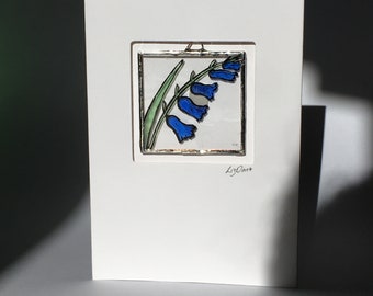 May include: A white card featuring a stained glass image of bluebells in shades of blue and green. The card is signed "LizOArt".