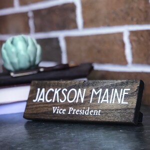 May include: A dark brown wooden desk nameplate with white text that reads "JACKSON MAINE Vice President".