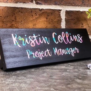 May include: Black wooden desk nameplate with the text "Kristin Collins Project Manager" in colorful script.