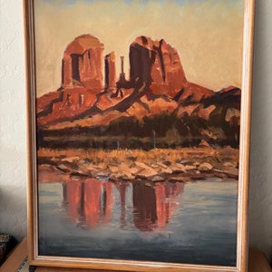 May include: Oil painting of a desert landscape with red rock formations reflected in a body of water. The artwork is framed in a light-colored wooden frame. The sky is a gradient of yellow and blue.