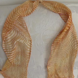 May include: A sheer, orange scarf with a white dotted pattern. The scarf is draped over a white patterned surface. The scarf is made of a lightweight material and is knotted at the top. The scarf is approximately 60 inches long.