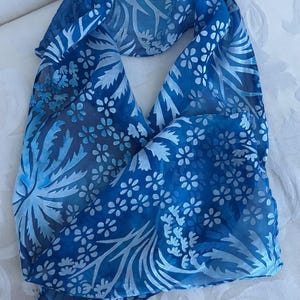 May include: A blue scarf with a floral pattern. The scarf features a design of white flowers and leaves against a blue background. The scarf is made of a lightweight, sheer fabric.